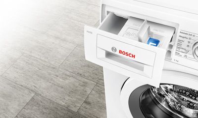 Das Bild zeigt einen modernen, zweiflügeligen Edelstahl-Kühlschrank, der offen steht und Lebensmittel sowie Getränke im Inneren sichtbar sind. Der Raum hat eine helle, freundliche Atmosphäre mit hellen Wänden und einer Pflanze auf einer Kommode im Hintergrund. Der Boden ist mit hellen Holzdielen ausgelegt.